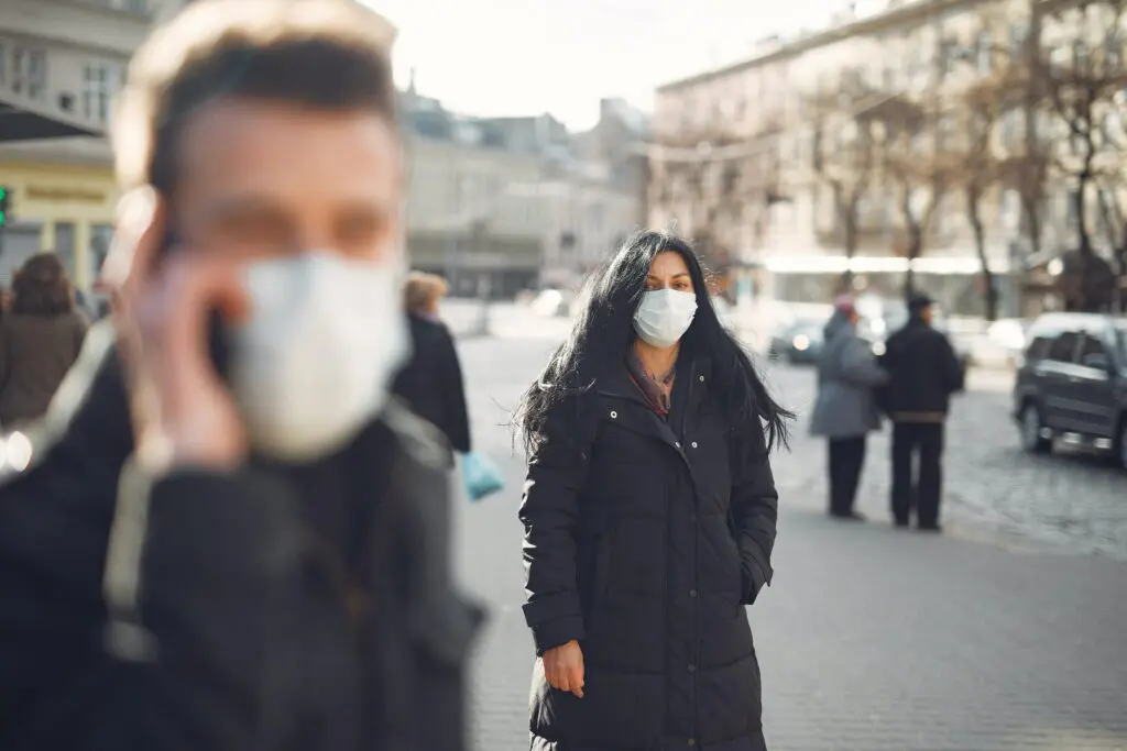 A group of people outside with masks on. The man in the foreground is blurry but you can see he's talking on a cell phone. The dark-haired woman behind him is looking toward the camera.