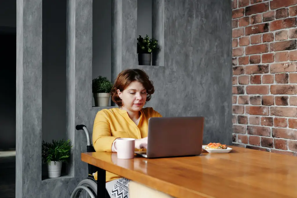 A woman in a yellow blouse sitting in a wheel chair at a table while using her laptop