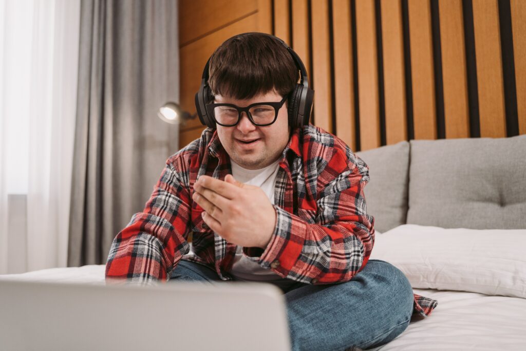 A young man with down syndrome sitting on a bed wearing headphones and using his laptop